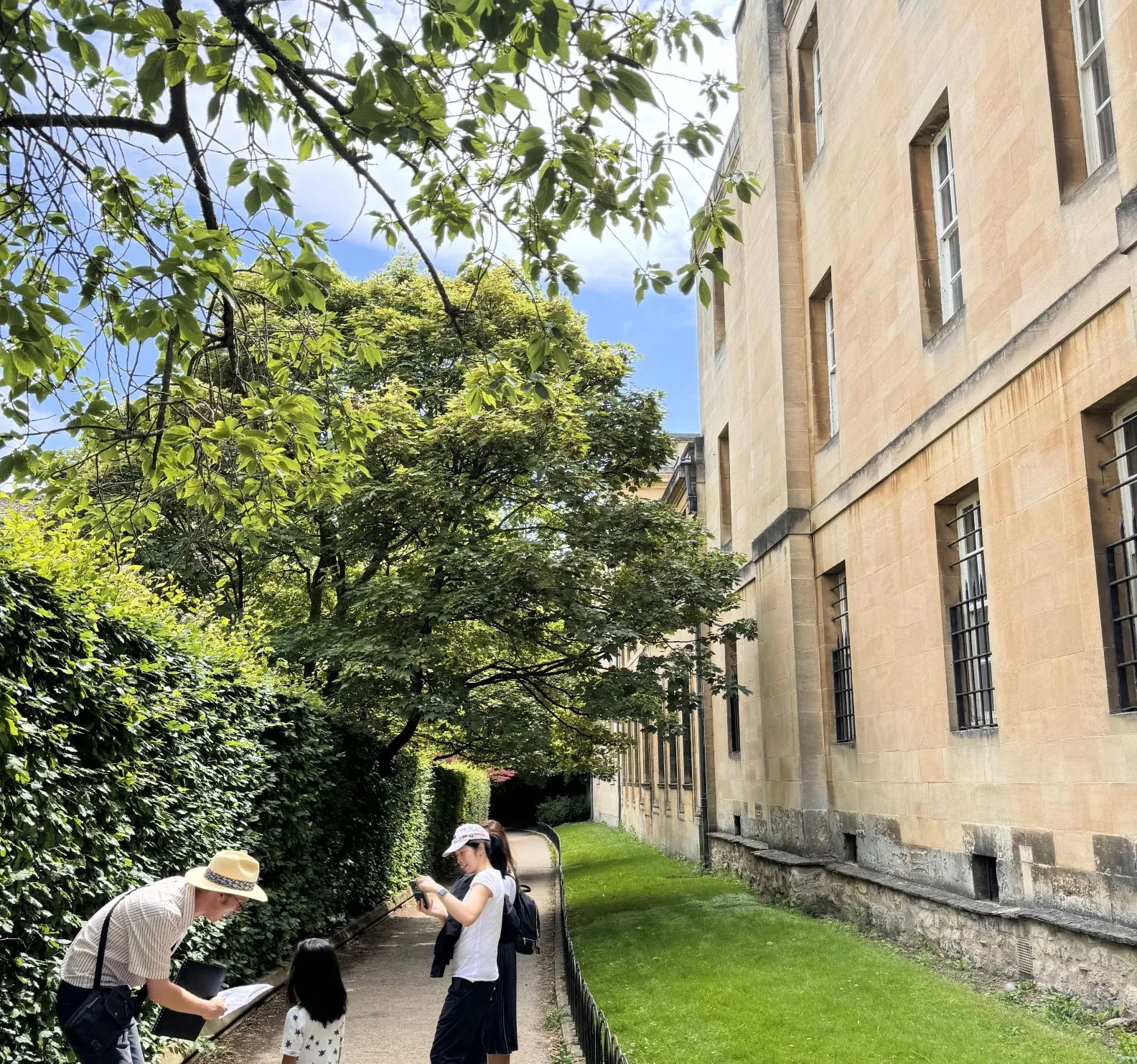 A tour guide explains a map to a young girl whilst her mother takes a photo. They are on a path flanked by a hedge and a stone building.